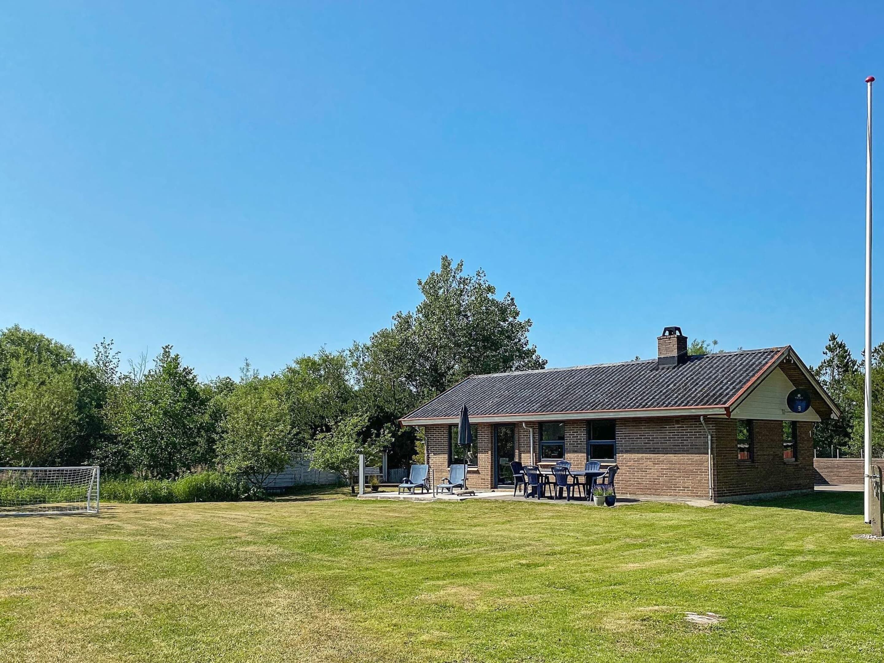 Ein kleines Backsteinhaus mit geneigtem Dach, einer Terrasse und Gartenmöbeln, vor einem strahlend blauen Himmel.