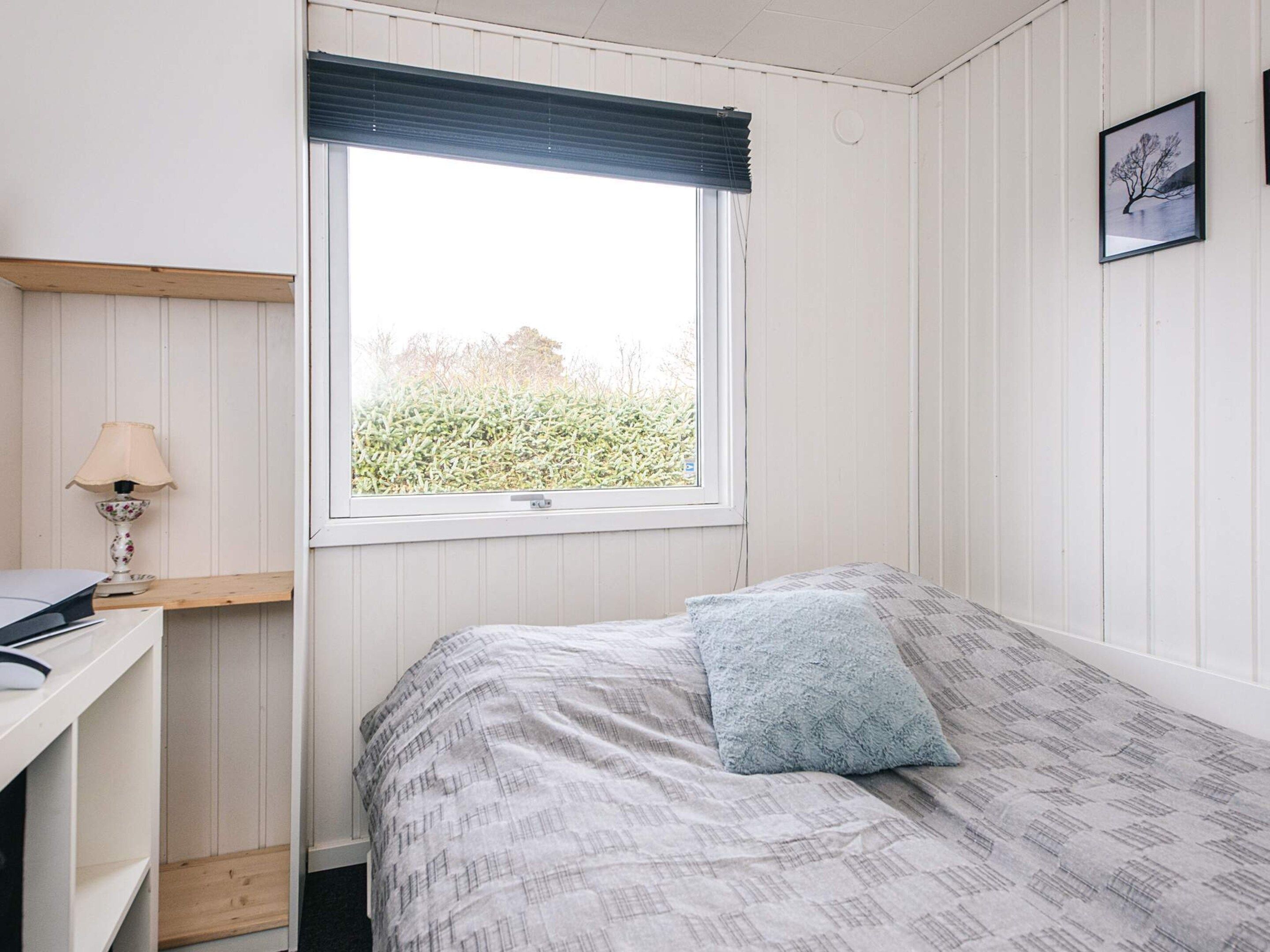 A bedroom with a bed, a desk, a lamp, a window with a view of greenery, and a framed picture on the wall.