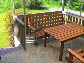 Et træbænk og et bord står på en stenbelagt terrasse med udsigt til en have.