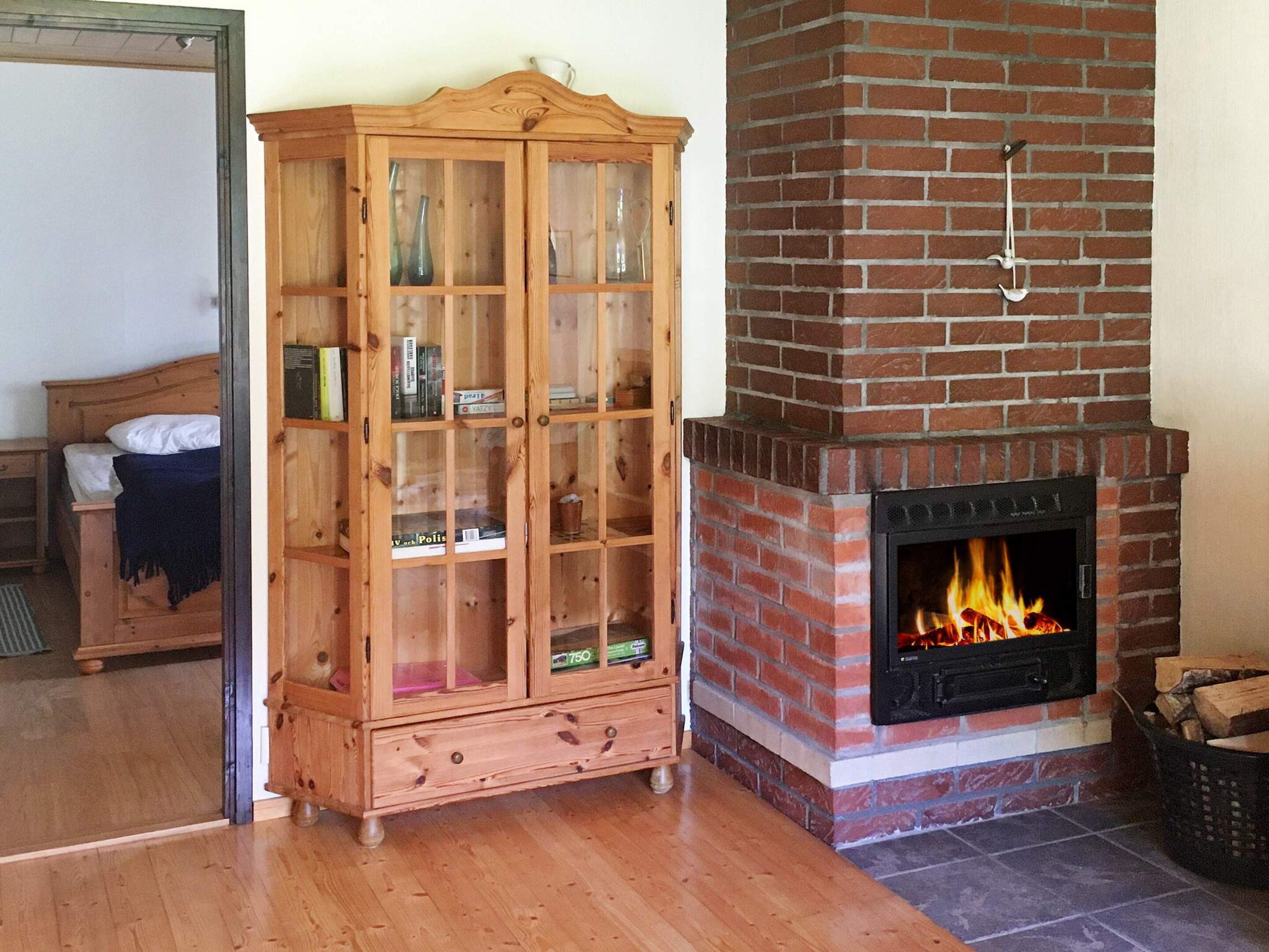A wooden cabinet with glass doors, a brick fireplace with a fire, and a bed in the background.