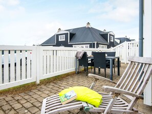 A patio area with a table and chairs, a white fence, and a building with a dark roof in the background.