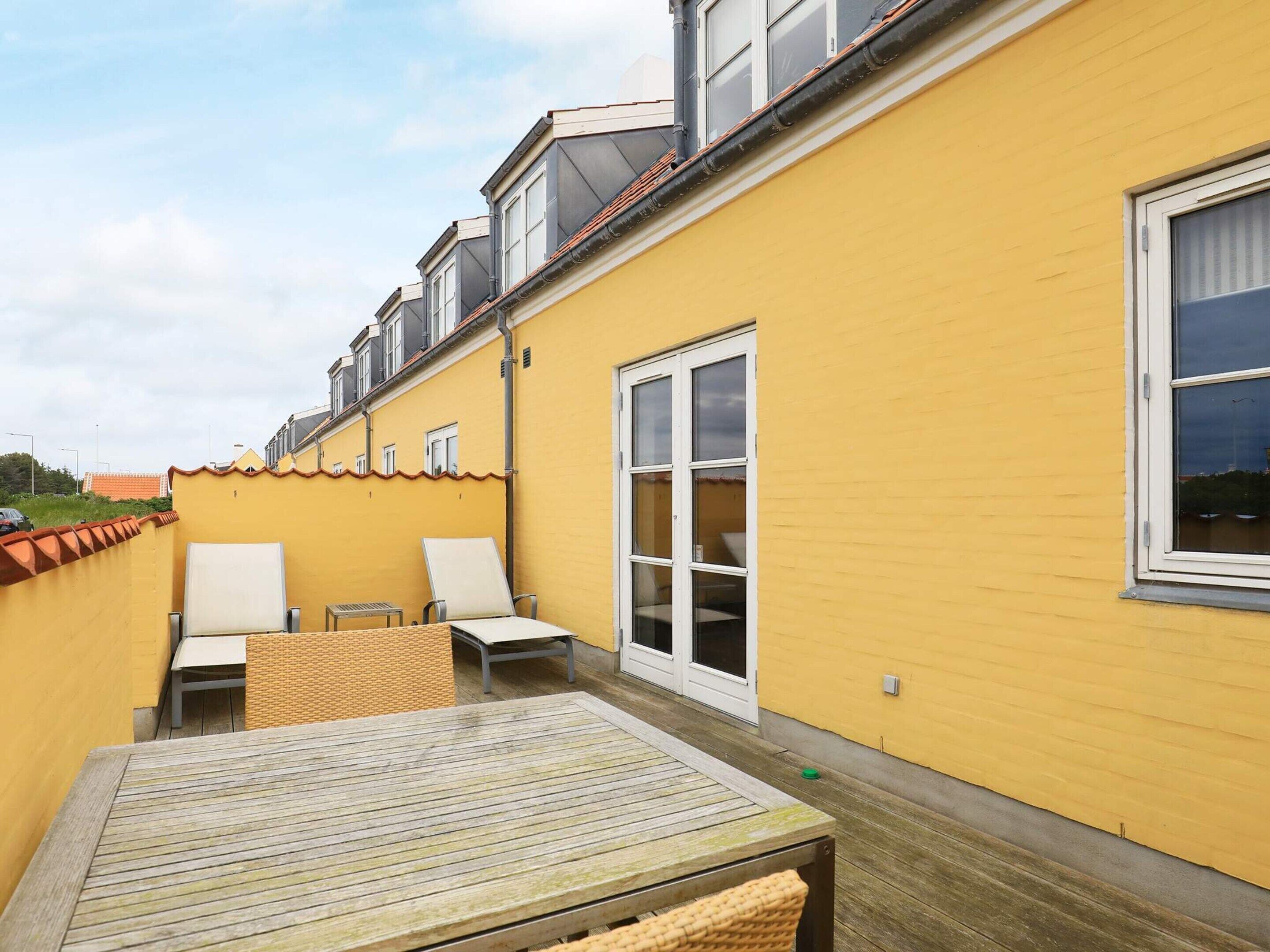 A yellow building with a wooden deck, two white chairs, and a table.