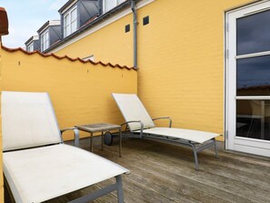 A balcony with two white lounge chairs and a small table, a yellow wall, and a window.