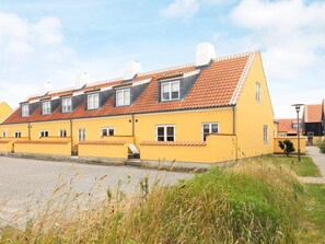 A yellow building with a sloped roof and multiple windows, located next to a paved area with grass and a lamp post.