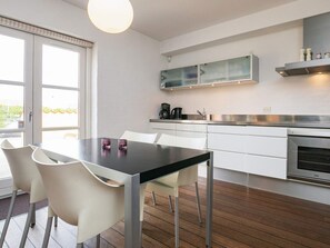 A modern kitchen with white cabinets, a black countertop, and a built-in oven. There is a dining table with chairs and a window with a view of greenery outside. - 4 Star Holiday Home in Skagen (Skagen)