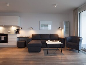 A modern living room with a sofa, a coffee table, and a chair. There is a kitchen area with cabinets and a coffee machine. - 5 Star Holiday Home in Wendtorf (Wendtorf)
