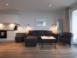 Una sala de estar moderna con un sofá, una mesa de centro y un sillón. Hay una zona de cocina con armarios y una cafetera.