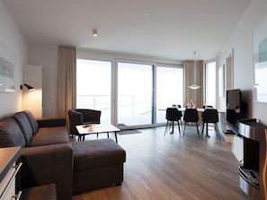 Quarto de hotel moderno com sofá, mesa de jantar, cadeiras, televisão de tela plana e varanda com vista.