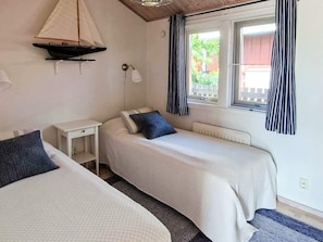 A bedroom with two beds, a small table, a wall-mounted lamp, and a window with curtains.