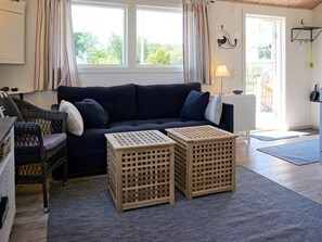 Ein gemütliches Wohnzimmer mit einem dunkelblauen Sofa, zwei Holz-Kisten, einem Korbstuhl und Blick ins Freie durch ein Fenster.