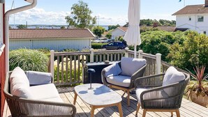 A wooden deck with outdoor furniture, including chairs and a table, overlooking a residential area with houses and a body of water in the background. - 4 Star Holiday Home in Onsala (Gottskar)