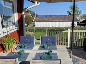 En uteplats med två blå sittstolar med kuddar, ett bord dukat med gröna tallrikar och muggar, en krukväxt och en utsikt över ett hus och vatten i bakgrunden.