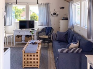 Un salon confortable avec un canapé, un téléviseur, un fauteuil en osier, une petite table et une vue sur l’extérieur grâce à de grandes fenêtres.