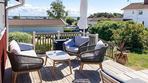 A wooden deck with outdoor furniture, including two wicker chairs and a small table, surrounded by greenery and a view of the sea. - 4 Star Holiday Home in Onsala (Gottskar)