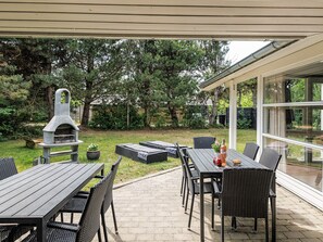 An outdoor patio with a stone barbecue, wicker furniture, and a glass-enclosed dining area. - 4 Star Holiday Home in Vaeggerlose (Vaeggerlose)