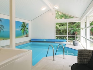 An indoor swimming pool with clear blue water, a blue cushioned bench, and a large window with a view of greenery. - 4 Star Holiday Home in Vaeggerlose (Vaeggerlose)