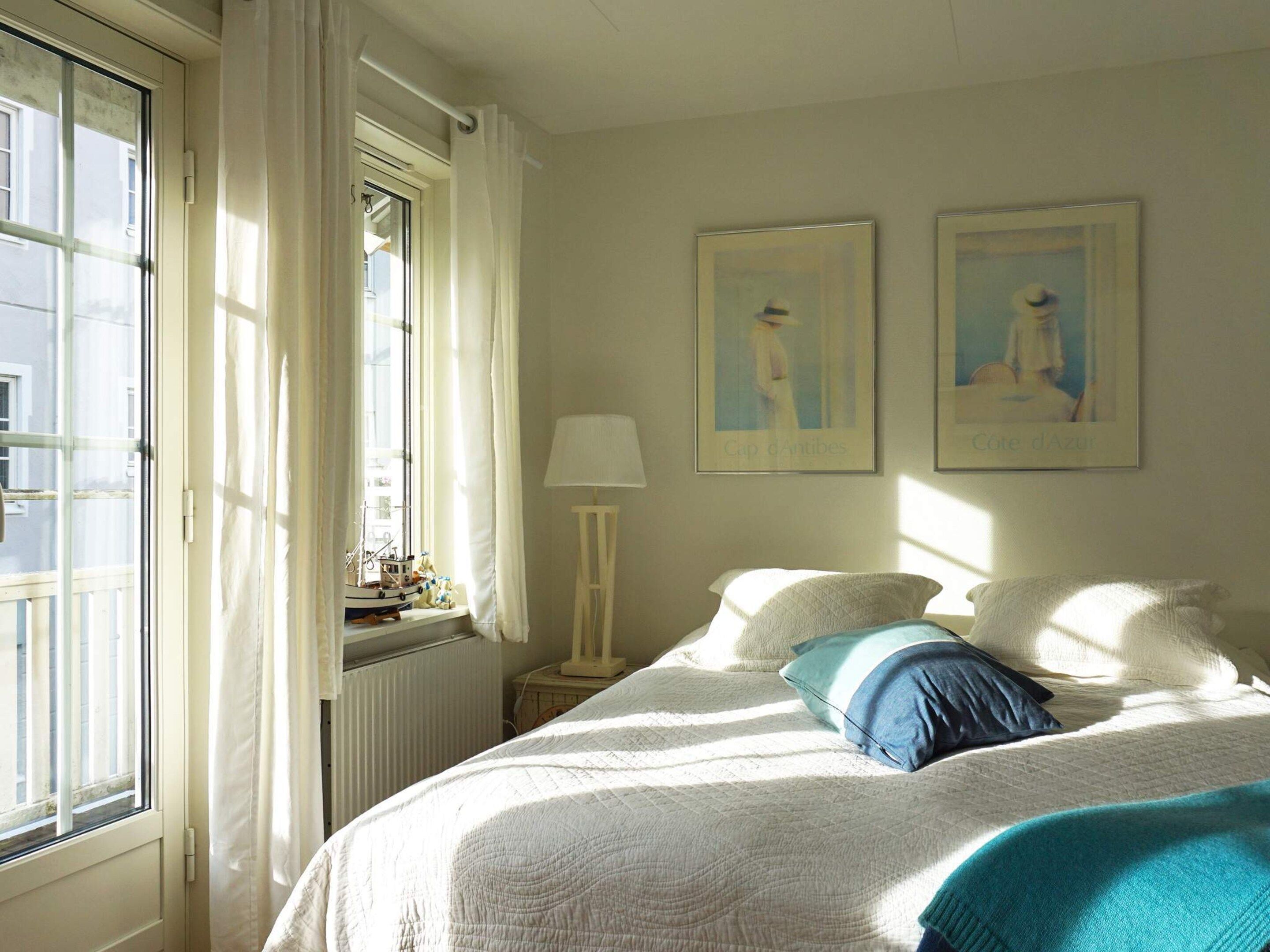 Ein Schlafzimmer mit einem Bett, einem Fenster mit Vorhängen, einem Heizkörper und zwei gerahmten Bildern an der Wand.