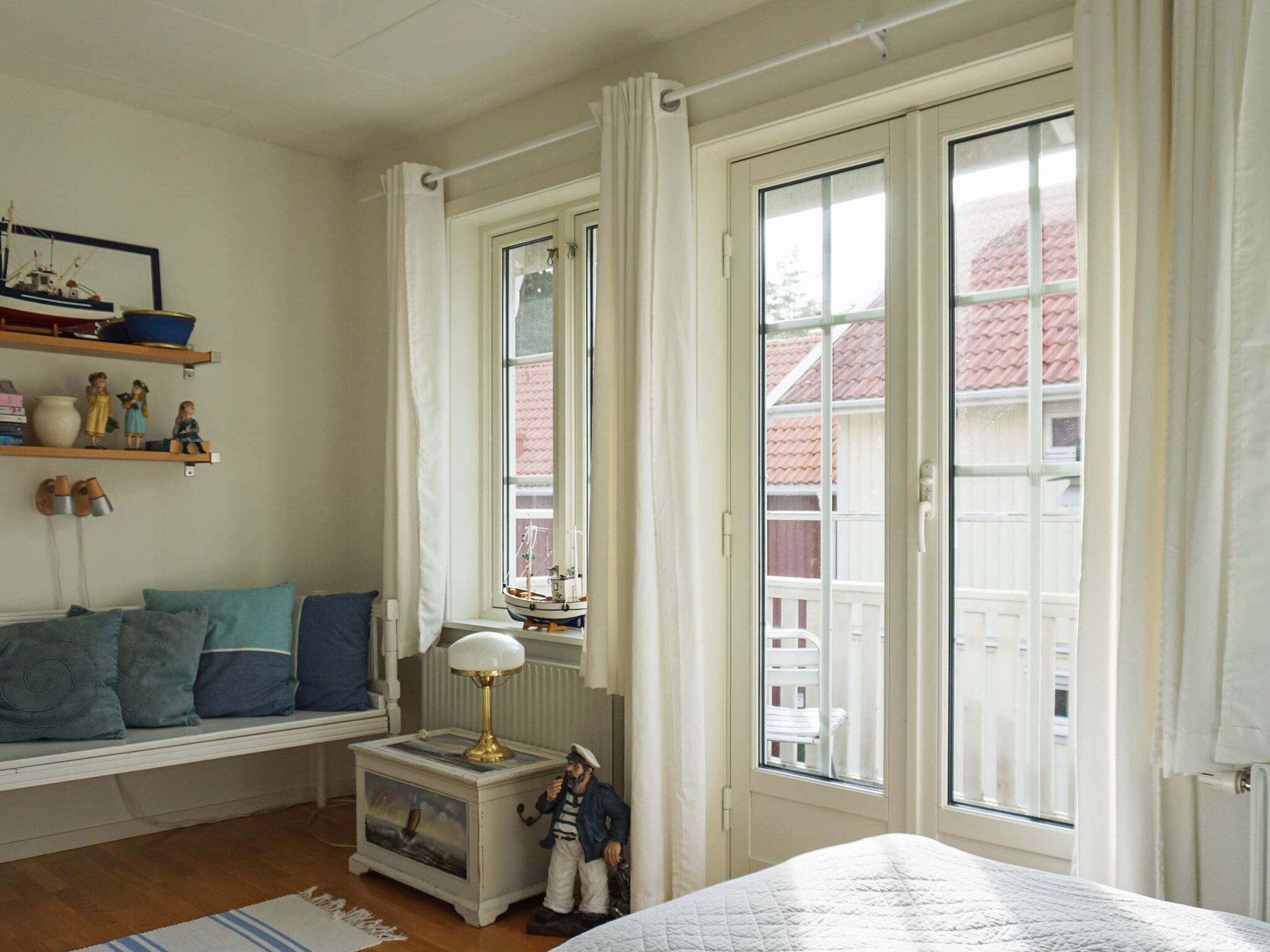 A bedroom with a bed, a nightstand with a lamp, a window with curtains, and a shelf with figurines.