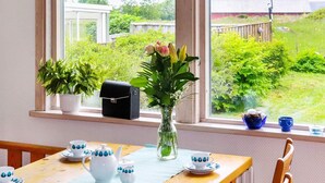 A dining area with a table set for tea, a vase with flowers, and a view of greenery and a building outside.