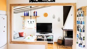 A cozy living room with a TV, wooden beams, a fireplace, and a kitchen area visible through an open doorway.