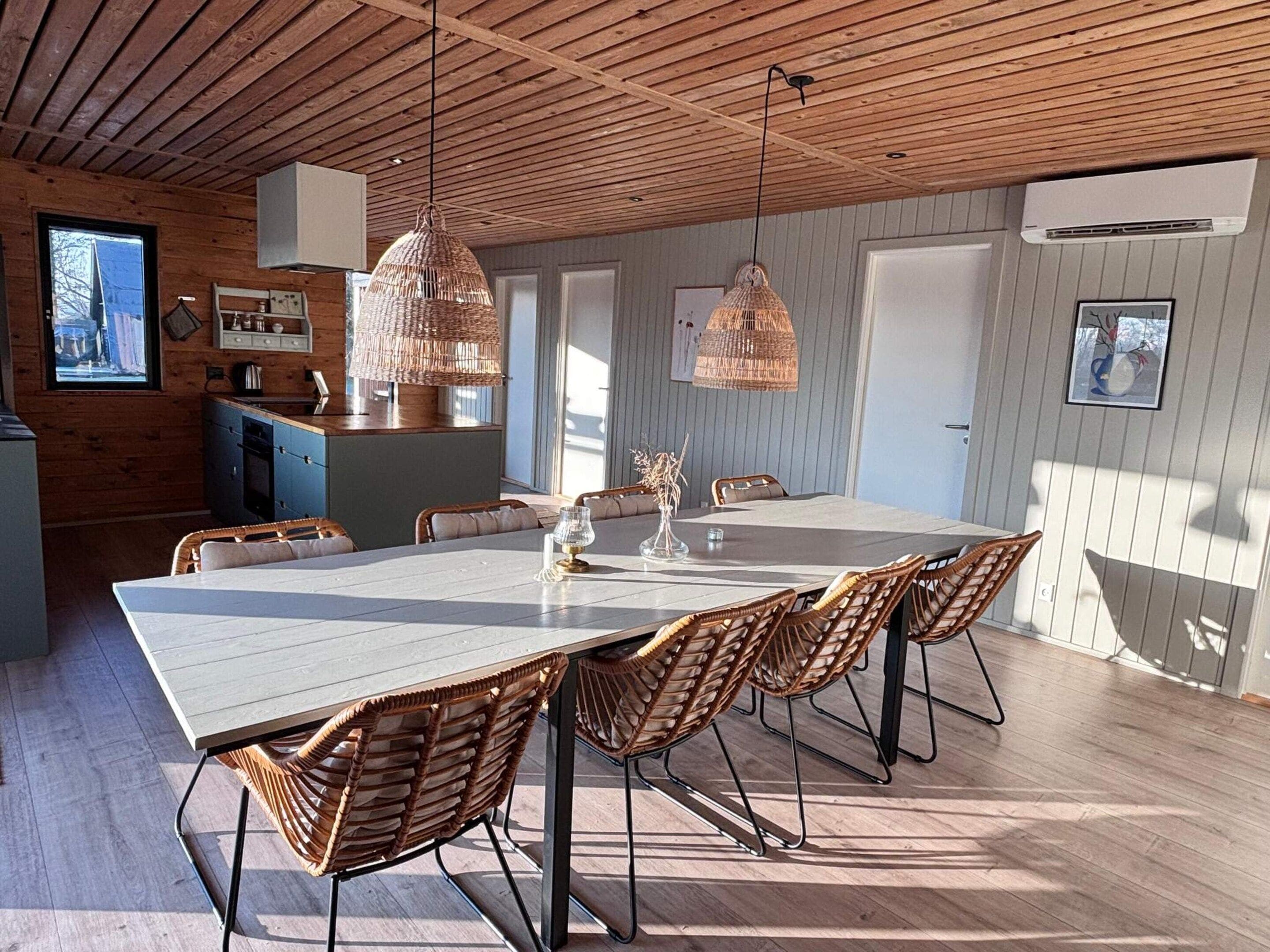 A spacious dining area with a long table and wicker chairs, a kitchen with wooden cabinets, and a hanging light fixture.