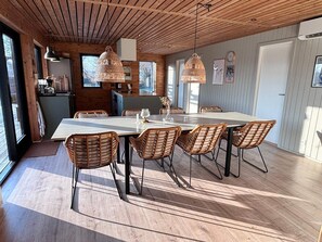 A dining area with a long table, wicker chairs, pendant lights, and a view through large windows.