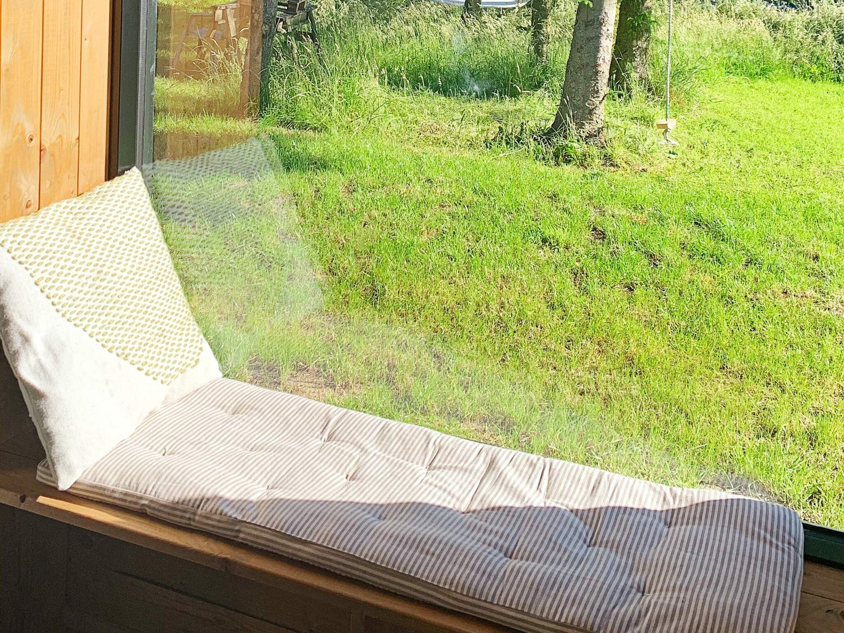 A bed with a striped mattress and a white pillow, positioned near a window overlooking a grassy area.