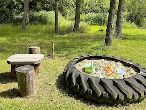 Un bac à sable rempli de pneus et de jouets, une table en bois et un tronc d’arbre en bois, le tout installé dans un espace extérieur recouvert d’herbe.