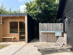 A wooden deck with a bench and a lounge chair, a wooden structure with a glass door, and a metal water feature.