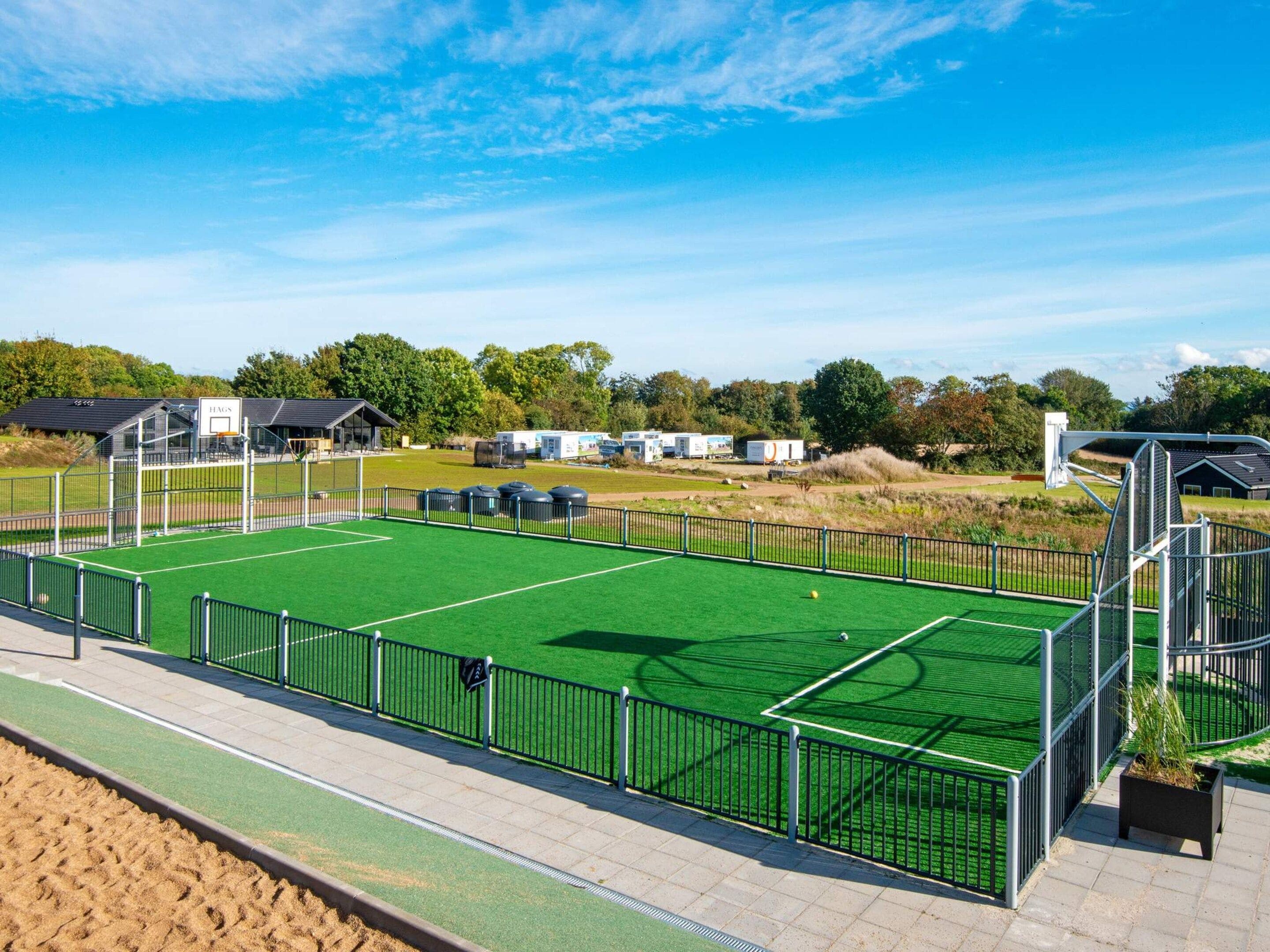 A sports court with a green surface, surrounded by a metal fence, and a building labeled 'HAG' in the background.