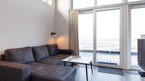 A modern living room with a sofa, a small table, and a lamp. There is a large sliding glass door leading to an outdoor balcony.