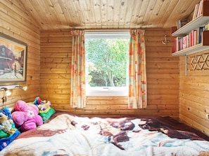 Acogedora habitación tipo cabaña de madera con una cama, peluches, una estantería y un ventanal con cortinas.