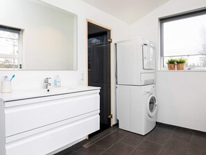 A modern bathroom with a white vanity, a built-in washing machine, a large mirror, and a window with plants. - 6 Person Holiday Home in Blokhus (Blokhus)