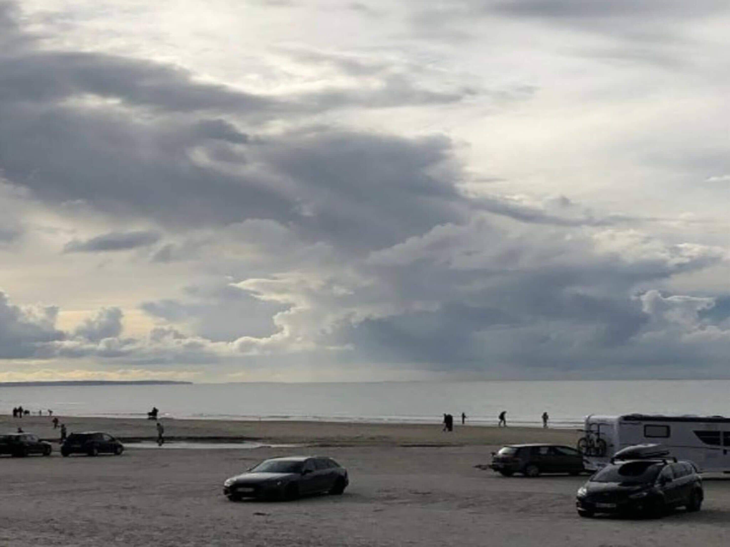 A beach with cars and a trailer, people walking, and a cloudy sky.