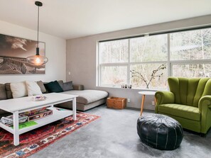 A modern living room with a sofa, coffee table, ottoman, and a large window with a view of trees. - 6 Person Holiday Home in Blokhus (Blokhus)