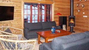A cozy log cabin interior with a sofa, wicker chairs, a wooden table, a TV, a fireplace, and a large window with red frames. - 4 Star Holiday Home in Salen (Salen)