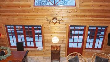 A log cabin interior with a dining area, two windows with red frames, a wall-mounted light fixture, and a decorative antler.