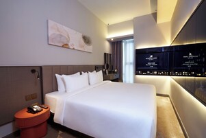 A modern hotel room with a large bed, a bedside table with a phone and remote, a wall-mounted artwork, and a digital display wall.