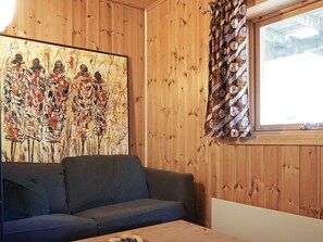 A room with a wooden paneled wall, a large abstract painting, a grey sofa, and a window with patterned curtains. - 6 Person Holiday Home in Stromstad (Stromstad)