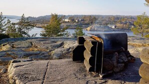 Un barbecue en pierre doté d’une cheminée, entouré de rochers et d’arbres, offrant une vue sur une étendue d’eau et des bâtiments situés au loin.