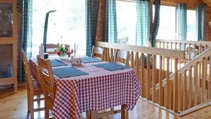 Interior de una cabaña de madera con una mesa de comedor lista para dos personas, sillas de madera y una escalera que sube a un nivel superior.