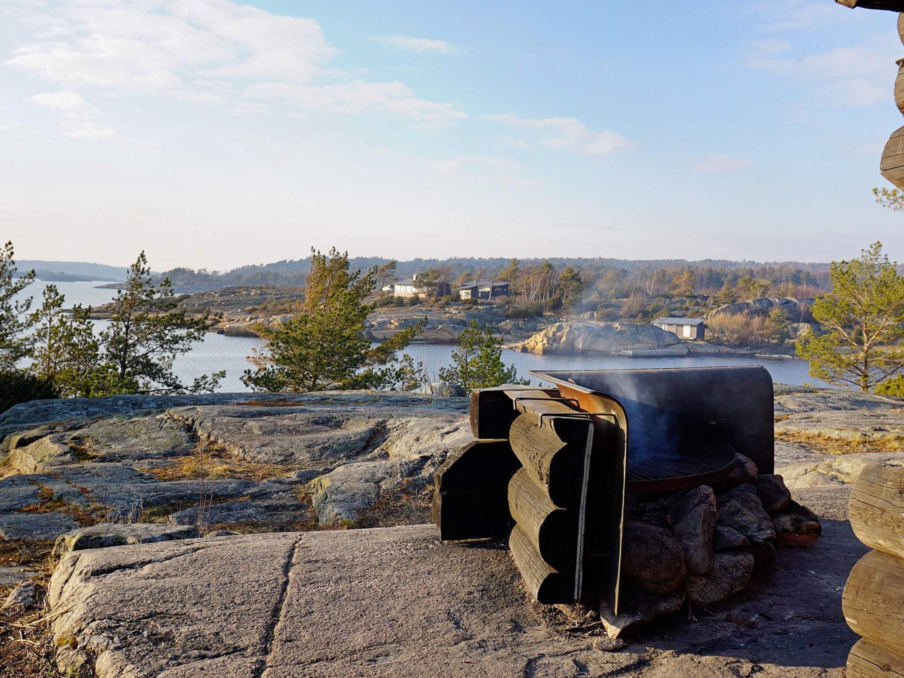 Una parrilla de piedra con chimenea, rodeada de rocas y árboles, con vistas a un cuerpo de agua y edificios al fondo.