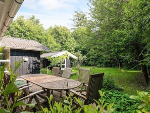 A wooden deck with a round table and chairs, surrounded by lush greenery and trees.
