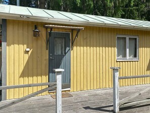 A yellow building with a dark door, a small window, and a wooden deck with railings. - 6 Person Holiday Home in Bottnaryd (Bottnaryd)