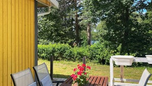 An outdoor wooden deck with a table and chairs, surrounded by greenery and a yellow building. - 6 Person Holiday Home in Bottnaryd (Bottnaryd)