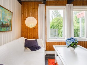 A room with a wooden wall, a white sofa, a round hanging light, a window with curtains, and a table with a flower vase.