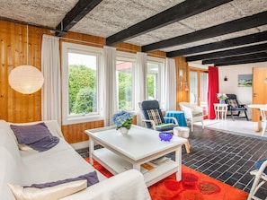 A cozy room with a white sofa, a small table with a vase, and a red rug. There are large windows with curtains, a wooden ceiling, and a door leading to another room.