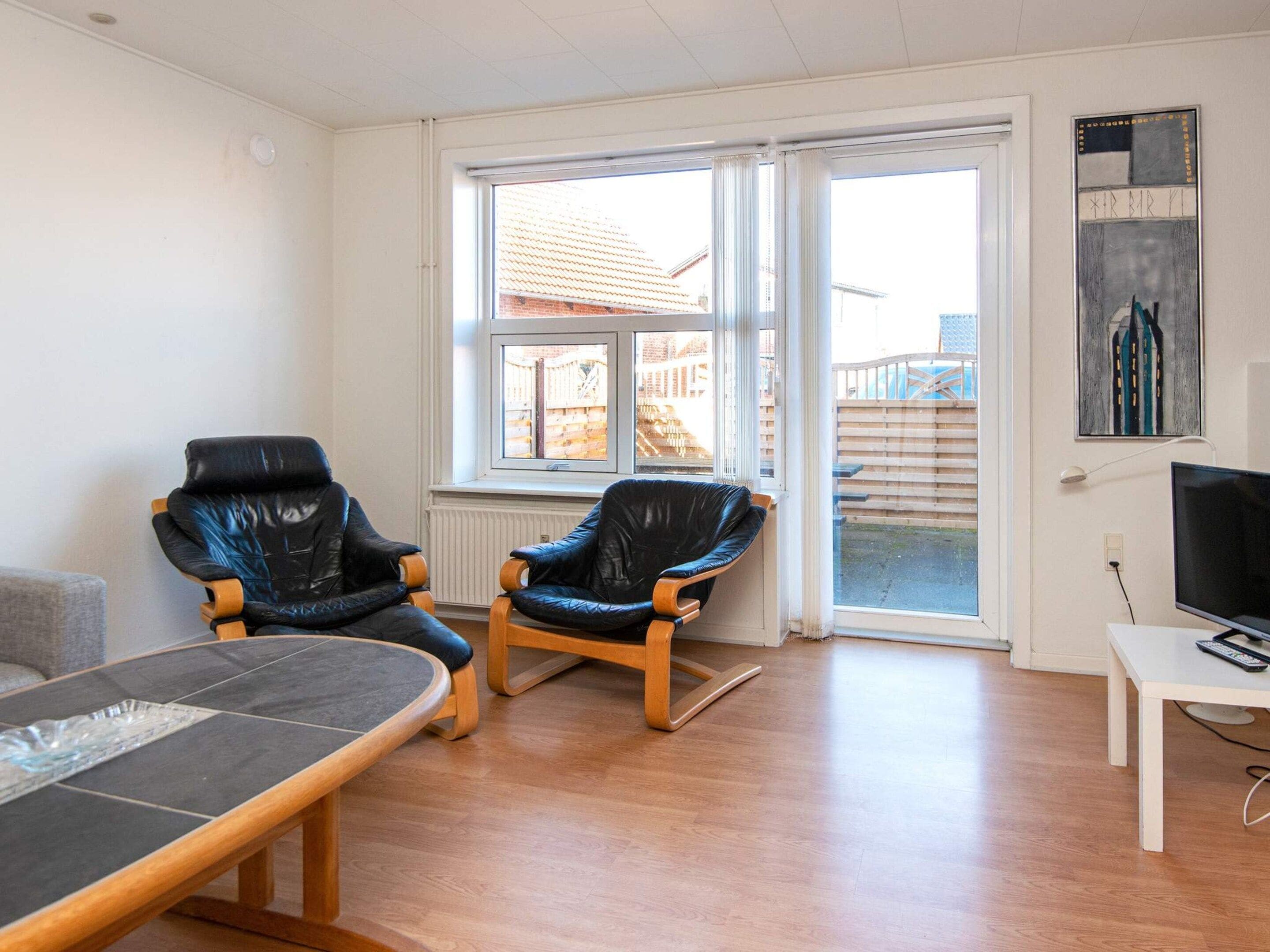 Ein Zimmer mit zwei Ledersesseln, einem Holztisch, einem Fernseher und einem Fenster mit Blick auf ein Gebäude und einen Zaun.