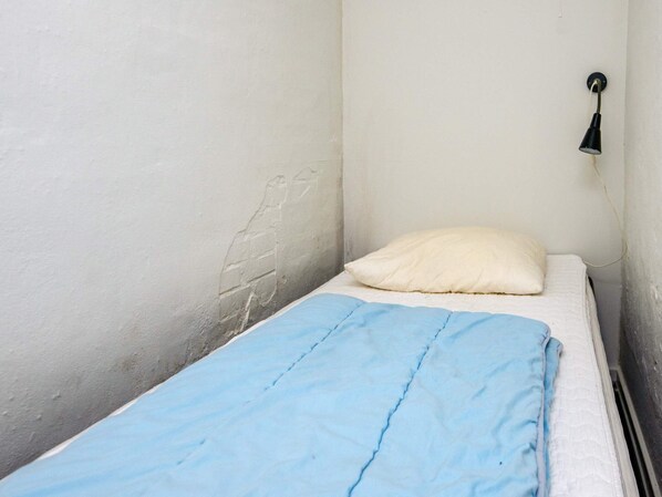 A small, single-bed room with a plain white wall and a single bedside lamp.