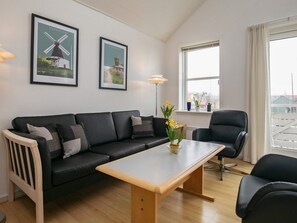 A modern living room with a sofa, a coffee table, and two armchairs. There are two framed pictures on the wall, a floor lamp, and a window with a view.